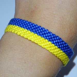 Puede incluir: Una pulsera tejida azul y amarilla, usada en la muñeca. La pulsera mide aproximadamente 2 cm de ancho y presenta un diseño a rayas, con azul en la parte superior y amarillo en la inferior.