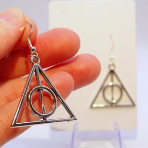 May include: Silver-colored Deathly Hallows earrings. The earrings feature a triangle with a circle and a vertical line through the center. The earrings are hanging from a silver hook. The earrings are displayed on a white background.