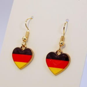 May include: A pair of heart-shaped earrings featuring the German flag design. The earrings have a gold-colored hook and a black, red, and yellow enamel finish. The earrings are displayed on a white background.