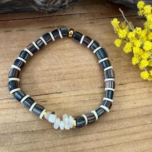 Peut inclure: Un bracelet de perles avec un mélange de perles bleu foncé, blanches et orange-rouge. Le bracelet comporte également une section de perles blanches et dorées. Le bracelet est présenté sur une surface en bois avec des fleurs jaunes en arrière-plan.