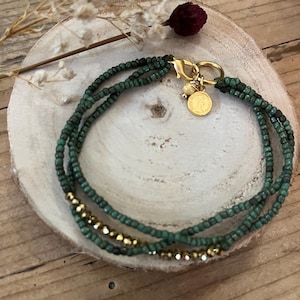 Peut inclure: Un bracelet en perles vertes et dorées avec un fermoir doré et un petit charme doré. Le bracelet est sur une surface en bois.