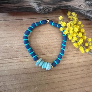 Peut inclure: Un bracelet fait à la main avec un mélange de perles bleues et turquoise. Le bracelet a une base bleu foncé avec des accents turquoise et une section de perles bleu clair de forme irrégulière. Des perles dorées séparent les sections. Le bracelet est présenté sur une surface en bois.