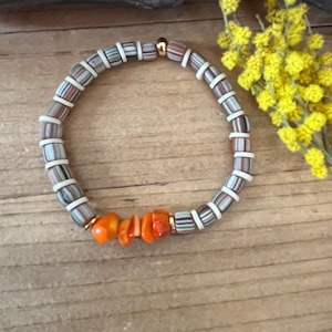 Peut inclure: Un bracelet de perles aux couleurs variées, notamment blanc, marron et gris, avec une perle dorée. Le bracelet comprend également un groupe de perles orange. Le bracelet est présenté sur une surface en bois.