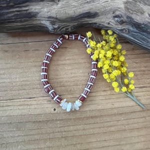 Peut inclure: Un bracelet de perles avec des perles rectangulaires bordeaux et blanches alternées. Le bracelet comprend une section de petites perles blanches de forme irrégulière et des accents dorés. Une branche de fleurs jaunes repose à proximité sur une surface en bois.
