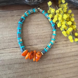 Peut inclure: Un bracelet de perles avec des perles turquoise, marron et dorées, et une section de pierres orange de forme irrégulière. Le bracelet est présenté sur une surface en bois. Des fleurs jaunes sont en arrière-plan. Circonférence du bracelet : environ 18 cm.