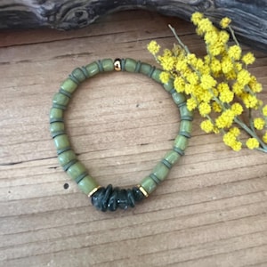 Peut inclure: Un bracelet de perles vertes avec des accents dorés et un groupe de pierres vert foncé. Le bracelet est présenté sur une surface en bois avec des fleurs jaunes et un morceau de bois flotté en arrière-plan.
