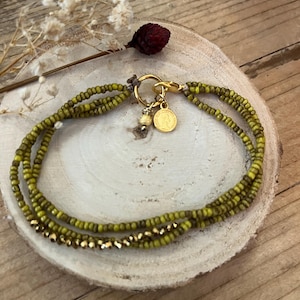 Peut inclure: Bracelet à plusieurs rangs de perles vertes et dorées. Le bracelet est composé de petites perles rondes et d'un fermoir doré avec un petit charme et un pendentif pièce. Présenté sur une surface en bois avec des fleurs séchées.