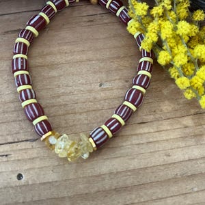 Peut inclure: Un bracelet de perles avec des perles rayées bordeaux et blanches, séparées par des perles jaunes. Il comporte également des cristaux jaune-orangé et des détails dorés. Une branche de fleurs jaunes est posée à côté, sur une surface en bois.