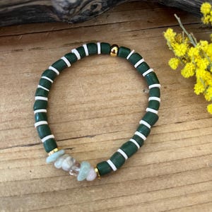 Peut inclure: Bracelet de perles vert foncé avec des accents blancs et des détails en métal doré. Le bracelet comporte un groupe de petites pierres de couleur claire. Le bracelet est présenté sur une surface en bois, avec des fleurs jaunes en arrière-plan.