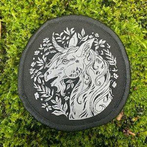 Pode incluir: Um remendo circular preto com um desenho bordado branco de uma cabeça de unicórnio com um fundo floral.