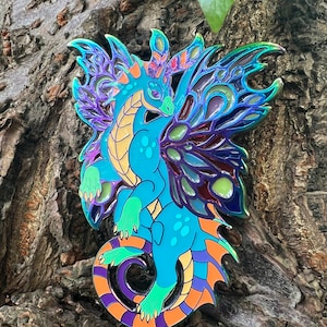 May include: An iridescent enamel pin of a blue and green dragon with wings that resemble stained glass. The dragon has orange and purple stripes on its tail.