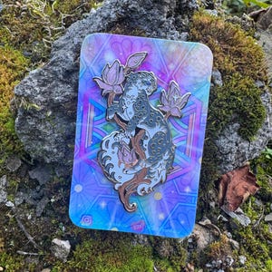 May include: A metal enamel pin featuring a stylised leopard perched on a branch surrounded by lotus flowers, set against a rectangular backing card with a vibrant, iridescent, geometric, and floral pattern in shades of blue, purple, and pink.