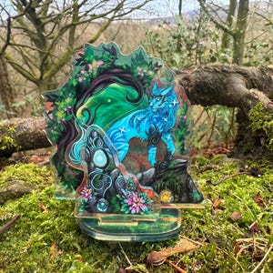 May include: A colorful acrylic stand featuring a blue and white wolf-like creature with star accents, set against a forest backdrop. The artwork includes green, pink, and blue hues, with floral and tree elements. The stand is displayed on a bed of green moss.