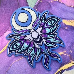 Peut inclure: Un patch brodé représentant un papillon de nuit avec une lune croissante derrière lui. Le papillon est bleu et violet avec des ailes détaillées et un corps blanc.