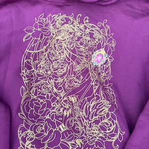 Puede incluir: Sudadera morada con un diseño floral dorado y una gema rosa. El diseño presenta la cara de una mujer rodeada de flores y hojas.