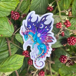May include: A blue and purple iridescent dragon with wings and a crescent moon tail charm. The dragon is on a white background with black and white polka dots.