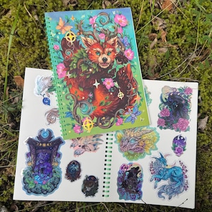 Puede incluir: Un cuaderno de espiral y una hoja de pegatinas con arte de fantasía. La portada del cuaderno muestra un zorro rojo con detalles florales y mariposas. La hoja de pegatinas incluye varias criaturas de fantasía en colores vibrantes, sobre un fondo de musgo.