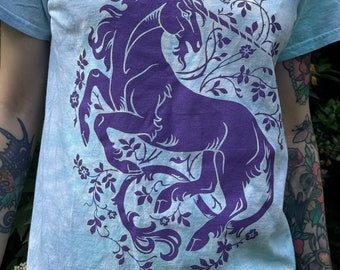 May include: A light blue t-shirt with a purple unicorn design. The unicorn is surrounded by a floral pattern.
