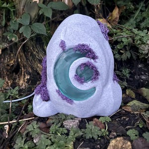 May include: A light gray, stone-like sculpture with a crescent moon-shaped opening. The moon is a translucent teal color. The sculpture is adorned with purple, moss-like material. The object is set in a natural outdoor environment.