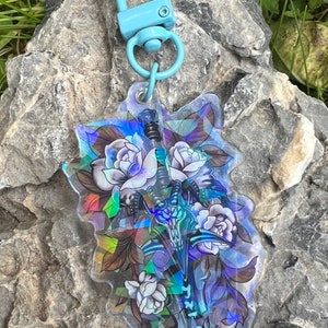 May include: A clear acrylic keychain with a holographic effect featuring a sword surrounded by white roses. The keychain has a light blue metal clasp.
