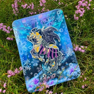 Könnte beinhalten: Ein Emaille-Pin mit einem Fantasy-Motiv, der ein geflügeltes Pferd mit gelb-schwarzer Mähne und lila gestreifter Rüstung zeigt. Der Pin ist vor einem blauen Hintergrund mit Schneeflocken und Blasen platziert.