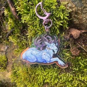 Puede incluir: Un llavero de acrílico transparente con un ciervo blanco con cuernos y un diseño floral morado y verde, con un cierre de metal plateado.