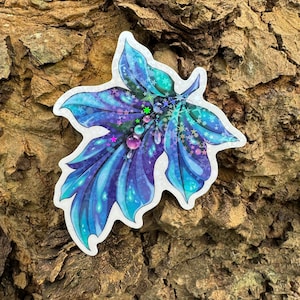 Puede incluir: Una pegatina iridiscente azul y púrpura de una hoja de arce con purpurina y gemas iridiscentes. La hoja está delineada en blanco y tiene un aspecto brillante y mágico.