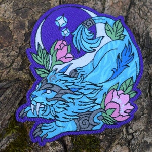 Könnte beinhalten: Ein gestickter Aufnäher mit einer mythischen Kreatur in Blau und Weiß, im Hintergrund ein Halbmond und Sterne. Die Kreatur hat einen grimmigen Ausdruck und ist von rosa Blumen und grünen Blättern umgeben.