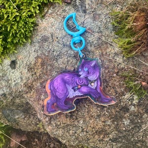 Puede incluir: Encanto de lobo morado con ojos azules y un diseño floral, unido a un llavero de plástico azul.