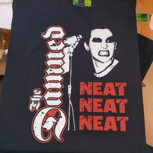 May include: Black t-shirt featuring a white graphic of a person singing into a microphone. The graphic also includes the text "The Damned" and "Neat Neat Neat" in red.
