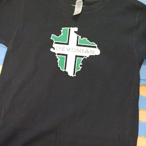 Pode incluir: Camiseta preta com um gráfico verde e branco da bandeira de Devon e a palavra "DEVONIAN" impressa nela.