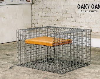 Puede incluir: Una moderna silla cuadrada de estructura de alambre con un cojín de cuero marrón. La silla es de metal oscuro y tiene un diseño minimalista. El logotipo de OAKY OAK furniture es visible en la esquina superior derecha.