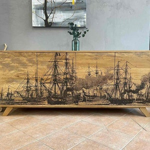 Puede incluir: Aparador de madera con cuatro puertas, con una ilustración en blanco y negro de veleros. El mueble tiene un acabado de madera natural y está sostenido por patas de madera en ángulo. La obra de arte representa una escena marítima histórica.