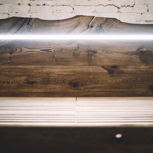 May include: A wooden bed frame with a headboard featuring a live edge design. The headboard is made of dark brown wood and has a built-in, horizontal LED light. The bed frame has a slatted base.