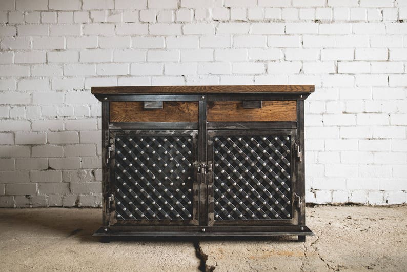 Puede incluir: Un mueble de estilo industrial con tapa de madera y dos cajones. Las puertas del mueble presentan un dise&ntilde;o de rombos con tachuelas met&aacute;licas. El marco es de metal oscuro y el mueble est&aacute; sobre un suelo de hormig&oacute;n. Mide unos 120 cm de ancho.