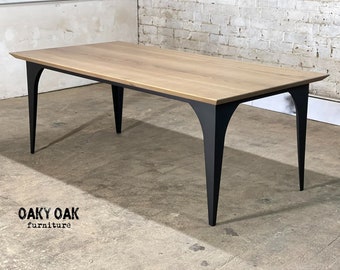 May include: A rectangular wooden dining table with a light oak finish. The table has a black metal frame and legs. The legs are curved and splayed. The table is in a room with a concrete floor and a white brick wall. The text "Oaky Oak furniture" is at the bottom.