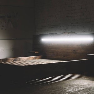 May include: A modern wooden bed frame with a built-in headboard featuring a horizontal, bright white light. The bed has a slatted base and small side tables. The wood has a natural grain.