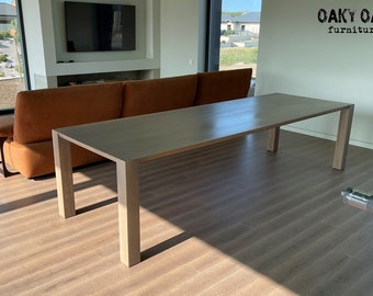 May include: A long, rectangular wooden dining table with a light finish. The table has a minimalist design with four square legs. The table is placed in a modern living room setting, with a brown sofa in the background. The logo "Oaky Oak furniture" is visible.