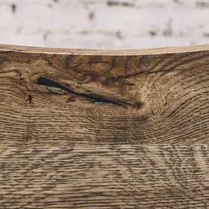 Puede incluir: Primer plano de una secci&oacute;n de madera envejecida, que muestra patrones de vetas naturales y un nudo. La madera tiene un tono marr&oacute;n c&aacute;lido con vetas m&aacute;s oscuras, lo que sugiere edad y textura. El fondo es una pared de ladrillos blancos borrosa.