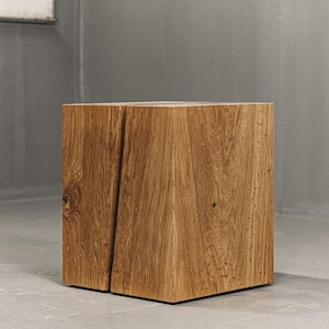 May include: A square-shaped wooden block stool featuring a natural light brown oak grain finish. The piece displays a prominent vertical fissure running down the front face, showcasing the raw texture and organic patterns of the wood material.
