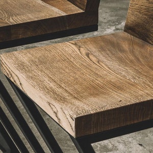 Puede incluir: Primer plano de dos sillas de madera con acabado de vetas naturales. Los asientos son rectangulares y est&aacute;n soportados por una estructura met&aacute;lica negra con un dise&ntilde;o geom&eacute;trico. Las sillas tienen una est&eacute;tica moderna y minimalista.