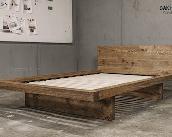 May include: A rustic wooden bed frame with a headboard and a low-profile design. The bed is made of light brown wood and features a slatted base. The bed frame is supported by thick, rectangular legs. The bed is in a room with a concrete floor and a gray wall.