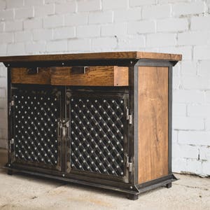 Puede incluir: Un mueble de madera con marco y detalles de metal oscuro. El mueble tiene dos cajones y dos puertas con un patr&oacute;n de diamantes. La madera es de color marr&oacute;n c&aacute;lido y el metal es gris oscuro. Es un mueble.
