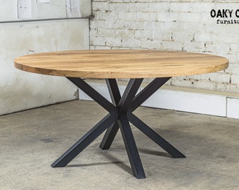 May include: A round wooden dining table with a light brown finish. The table has a black metal starburst base. The table is in a room with a white brick wall and a concrete floor. The text "Oaky Oak furniture" is visible.