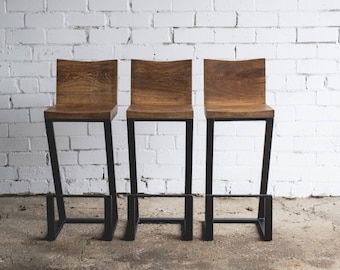 Puede incluir: Tres taburetes de bar de madera con asientos marrones oscuros y marcos de metal negro. Los asientos tienen un diseño curvo y las patas están en ángulo. Los taburetes están colocados contra una pared de ladrillos blancos.