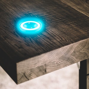 Puede incluir: Primer plano de un escritorio de madera con una luz azul brillante incorporada. El escritorio tiene un acabado de vetas de madera oscura y texturizada. La luz es circular y está incrustada en la superficie del escritorio.