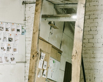 May include: A large, rectangular full-length mirror with a natural wood frame. The frame has a raw, unfinished edge and is supported by a black metal stand. The mirror is leaning against a white brick wall in a workshop setting.
