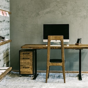 May include: A modern home office setup featuring a wooden desk with a computer monitor, a wooden chair, and a rolling file cabinet. The desk is supported by a black metal leg. The room has a gray wall and wooden shelves.