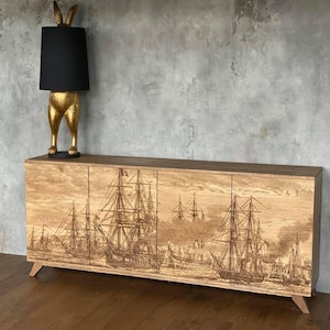 Puede incluir: Un mueble de madera con cuatro puertas, con una escena náutica grabada de veleros. Una lámpara dorada con forma de conejo y pantalla negra está encima. El mueble tiene patas anguladas y está contra una pared gris.