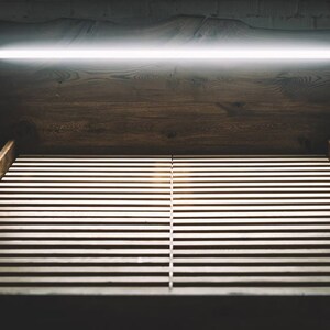 May include: A dark, moody image of a wooden bed frame with a slatted base. Above the bed, a wooden headboard and a bright, linear light fixture are visible. The scene is dimly lit, creating a sense of calm.